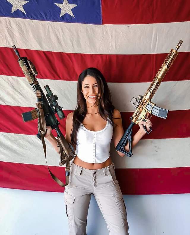 Luna souriante, tenant un fusil dans chaque main devant un drapeau américain