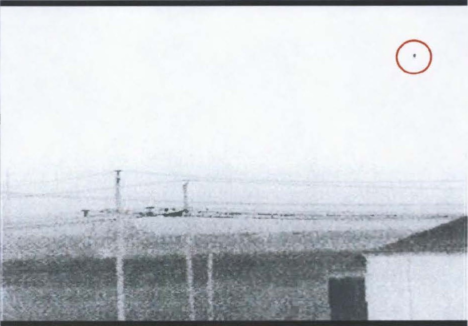 Image grise d'une caméra de surveillance montrant au loin des poteaux et un bâtiment, avec un point sombre entouré de rouge dans le ciel